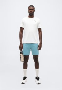 Άνδρας που στέκεται φορώντας λευκό t-shirt Puma, μπλε σορτς, λευκές κάλτσες και αθλητικά παπούτσια, κρατώντας ένα μπουκάλι νερό στο δεξί του χέρι.