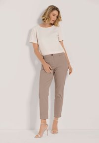 Beige blouse met korte mouwen gecombineerd met lichtbruine tapered pants. Het model draagt sandalen met blote voeten; de pants hebben een knoop aan de voorkant en zakken.