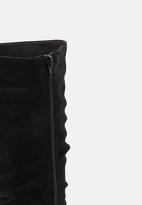 Bottine en daim noir avec un design texturé et ondulé, et une fermeture éclair latérale. L'intérieur semble doux et lisse, complétant la texture extérieure.