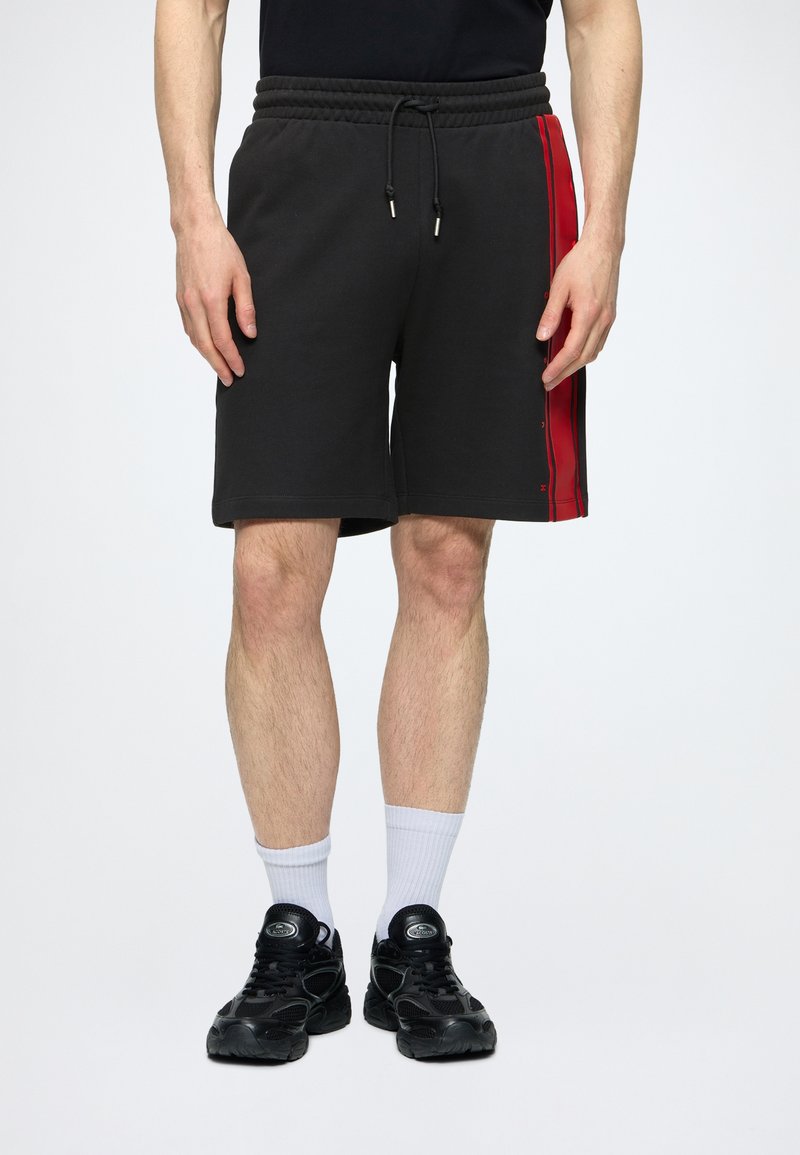 Homme portant un short noir avec des bandes rouges sur les côtés, des chaussettes blanches montantes et des baskets noires, debout devant un fond clair uni.