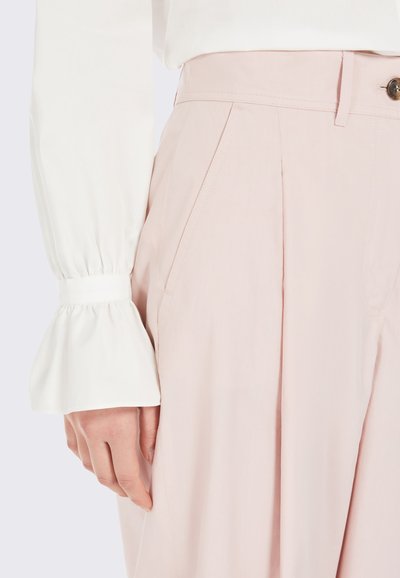 Pantaloni su misura rosa chiaro con pieghe e tasche laterali, abbinati a una blusa bianca a maniche lunghe con polsini a campana.