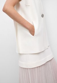 Haut sans manches blanc avec une texture lisse et des poches latérales, superposé à une jupe plissée légère en rose doux. Détail de boutons métalliques ronds.