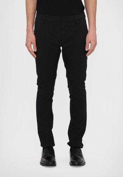 Schwarze Slim-Fit-Hosen mit glatter Textur, ausgestattet mit Gürtelschlaufen und Reißverschluss. Kombiniert mit glänzenden schwarzen Schuhen.