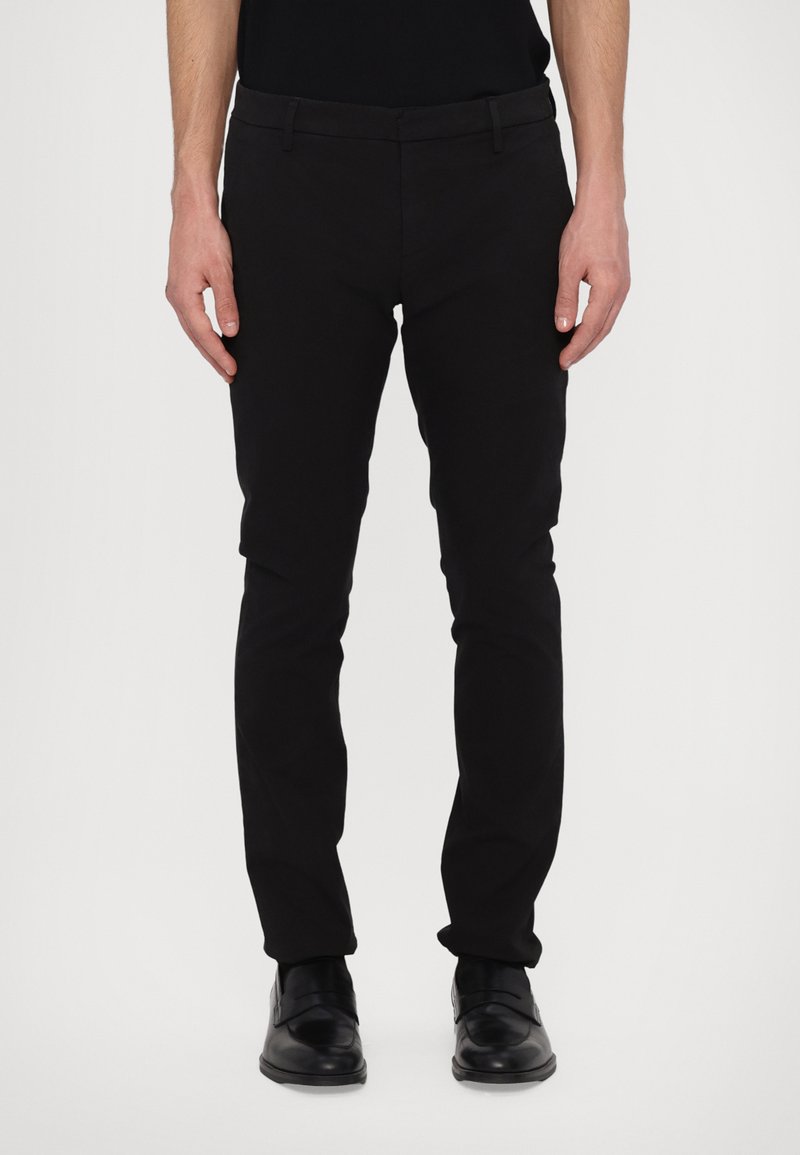 Svart slim-fit byxor med en mjuk textur, utrustade med bälteshällor och dragkedja. I kombination med polerade svarta skor.