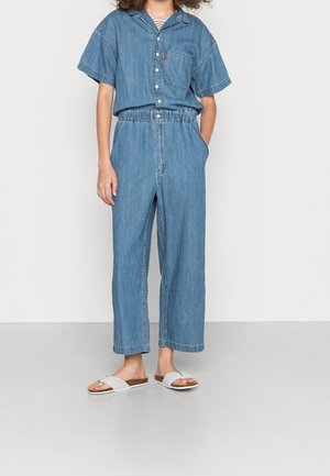 Personne portant une chemise en denim à manches courtes rentrée dans un pantalon en denim ample taille haute et des sandales blanches à enfiler, debout devant un fond uni.
