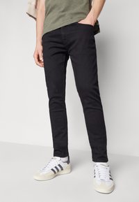 Persona vestida con pantalones negros ajustados, camisa verde oliva y zapatillas blancas con rayas negras, de pie con una mano en el bolsillo.