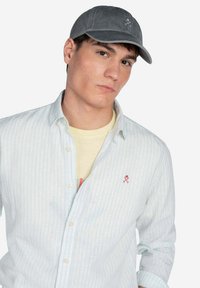 Gorra gris con visera curva, hecha de tela suave; adornada con un simple logo blanco. Camisa de botones a rayas claras llevada sobre una camiseta amarilla.