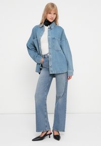 Giacca di denim blu chiaro con grandi tasche, abbinata a jeans a vita alta blu chiaro a gamba larga. Le mules nere con tacco completano l'outfit.