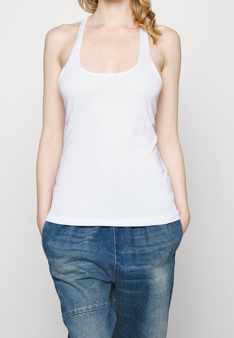 Femme portant un débardeur blanc sans manches et un jean bleu ample, les mains dans les poches, sur un fond clair uni.