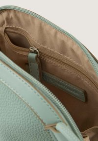 Zielona miętowa skórzana torba od wewnątrz posiada beżowe wykończenie, zawiera kieszeń na zamek oraz dekoracyjną metkę, prezentując teksturowaną powierzchnię z kontrastowymi elementami metalowymi.