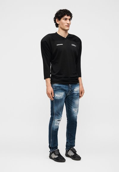 Νέος άνδρας με σγουρά μαλλιά που φοράει μακρυμάνικη μαύρη μπλούζα με κείμενο, φθαρμένα μπλε τζιν και μαύρα αθλητικά παπούτσια με λευκές ρίγες, όρθιος.