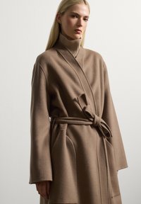 Cappotto lungo stile avvolgente in morbido tessuto marrone con vita con cintura, colletto a dolcevita a coste e tasche laterali. Texture liscia e vestibilità rilassata.
