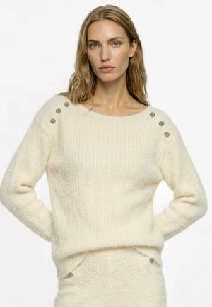 Kvinde med langt blond hår iført en flødefarvet, blød sweater med metalliske knapper på skuldrene og matchende nederdel, stående med hænderne på hofterne.