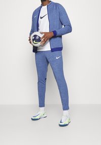 Persoon in een blauwe Nike trainingspak en een wit shirt staat met een witte en blauwe Nike voetbal tegen een effen achtergrond.