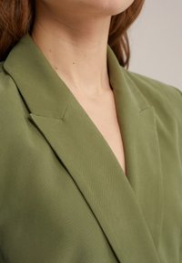 Olivově zelený blazer s hladkým, texturovaným materiálem, s širokými klopami a hlubokým výstřihem do V. Detailní záběr na výstřih a límec.