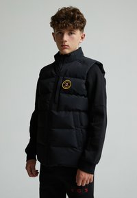 Gilet imbottito nero con colletto alto, chiusura con zip, tasche laterali e un patch rotondo sul petto. Indossato sopra una maglietta nera a maniche lunghe.