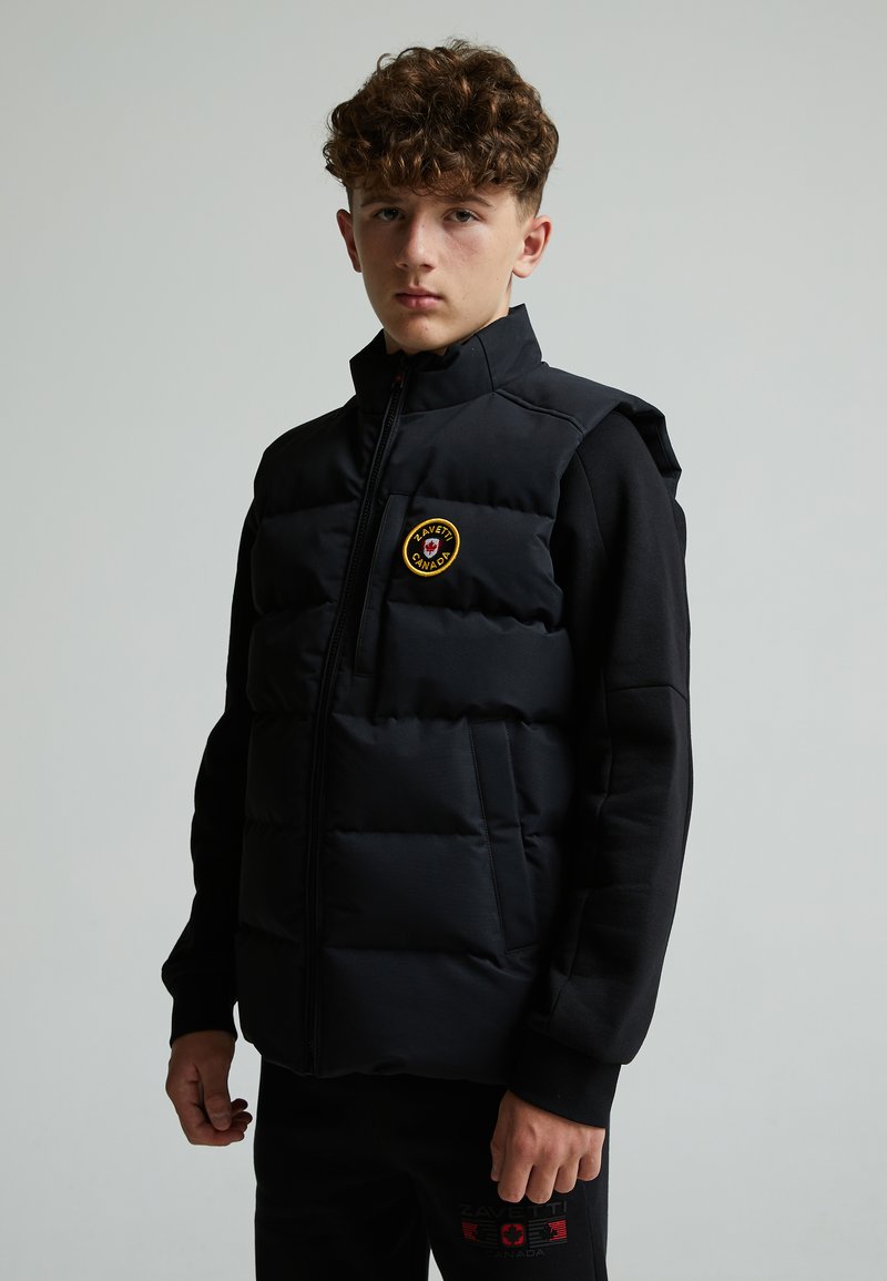 Gilet imbottito nero con colletto alto, chiusura con zip, tasche laterali e un patch rotondo sul petto. Indossato sopra una maglietta nera a maniche lunghe.