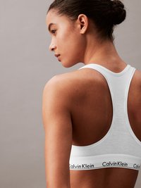 White cotton sports bra featuring a racerback design and a black Calvin Klein logo band at the bottom. Smooth texture and fitted shape.