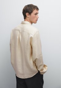Camicia in lino a maniche lunghe di un leggero beige, con colletto, polsini abbottonati e una vestibilità leggermente loose, mostrata da dietro.