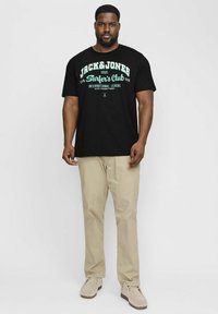 Schwarzes Baumwoll-T-Shirt mit weißem und tealfarbenem Text, kombiniert mit hellbeigen Hosen und beigen Wildlederschuhen. Einfaches Design mit lockerer Passform.