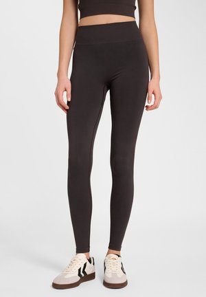 Vrouw draagt zwarte leggings met hoge taille en witte sneakers, staand tegen een eenvoudige lichte achtergrond.