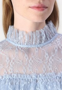 Top in pizzo azzurro chiaro con colletto alto e arricciato, motivi floreali intricati e una texture trasparente, con una finitura morbida e delicata.
