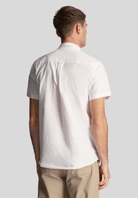Camicia bianca a maniche corte realizzata in tessuto morbido. Presenta un colletto classico e un rinforzo posteriore. L'orlo è leggermente curvato, con polsini arrotolati e senza motivi visibili.