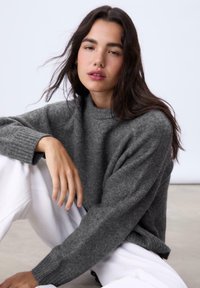 Grauer Strickpullover mit hohem Kragen und gerippten Bündchen, kombiniert mit weißen Hosen, die eine weiche Textur und einen lockeren Schnitt aufweisen.