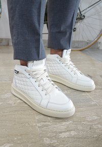 Sneaker alte bianche in pelle, con un motivo trapuntato, lacci beige e una suola di colore chiaro. Indossate con pantaloni grigi.