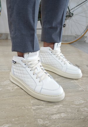 Weiße High-Top-Sneakers aus Leder mit einem gesteppten Muster, beigen Schnürsenkeln und einer hellen Sohle. Getragen mit grauen Hosen.