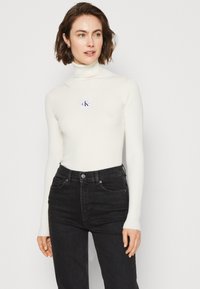 Maglione a collo alto bianco a costine con maniche lunghe, caratterizzato da una piccola patch con il logo CK, abbinato a jeans neri a vita alta.