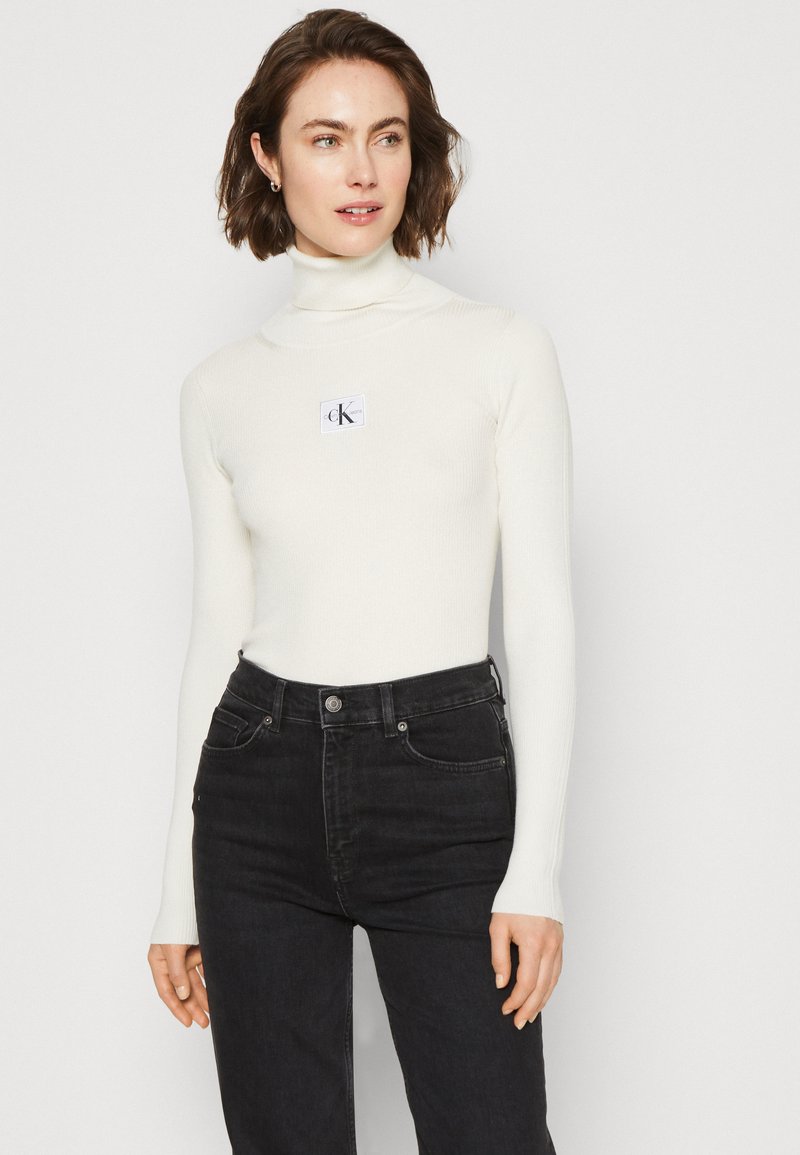 Maglione a collo alto bianco a costine con maniche lunghe, caratterizzato da una piccola patch con il logo CK, abbinato a jeans neri a vita alta.