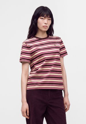 Young woman with long black hair wearing a short-sleeve striped t-shirt in shades of brown and cream, paired with dark pants against a plain background.