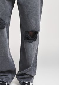 Μαύρο τζιν με σκισίματα και μεγάλα φθαρμένα ανοίγματα στα γόνατα, φορεμένο με μαύρα και λευκά αθλητικά παπούτσια μπροστά σε απλή φόντο.