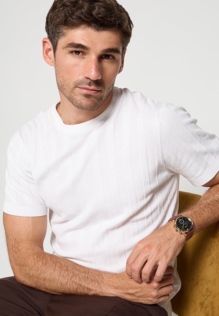 Camiseta blanca de punto de manga corta con textura de canalé vertical. La persona lleva un reloj de pulsera con cara negra y correa de cuero marrón.