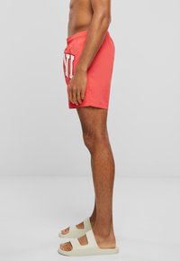 Red swimming shorts with white logo detail, elastic waistband, and a loose fit. Paired with light-colored slide sandals.