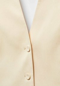 Blouse beige clair avec un col en V, ornée de deux boutons ronds à l'avant. Le tissu semble lisse avec un léger éclat.