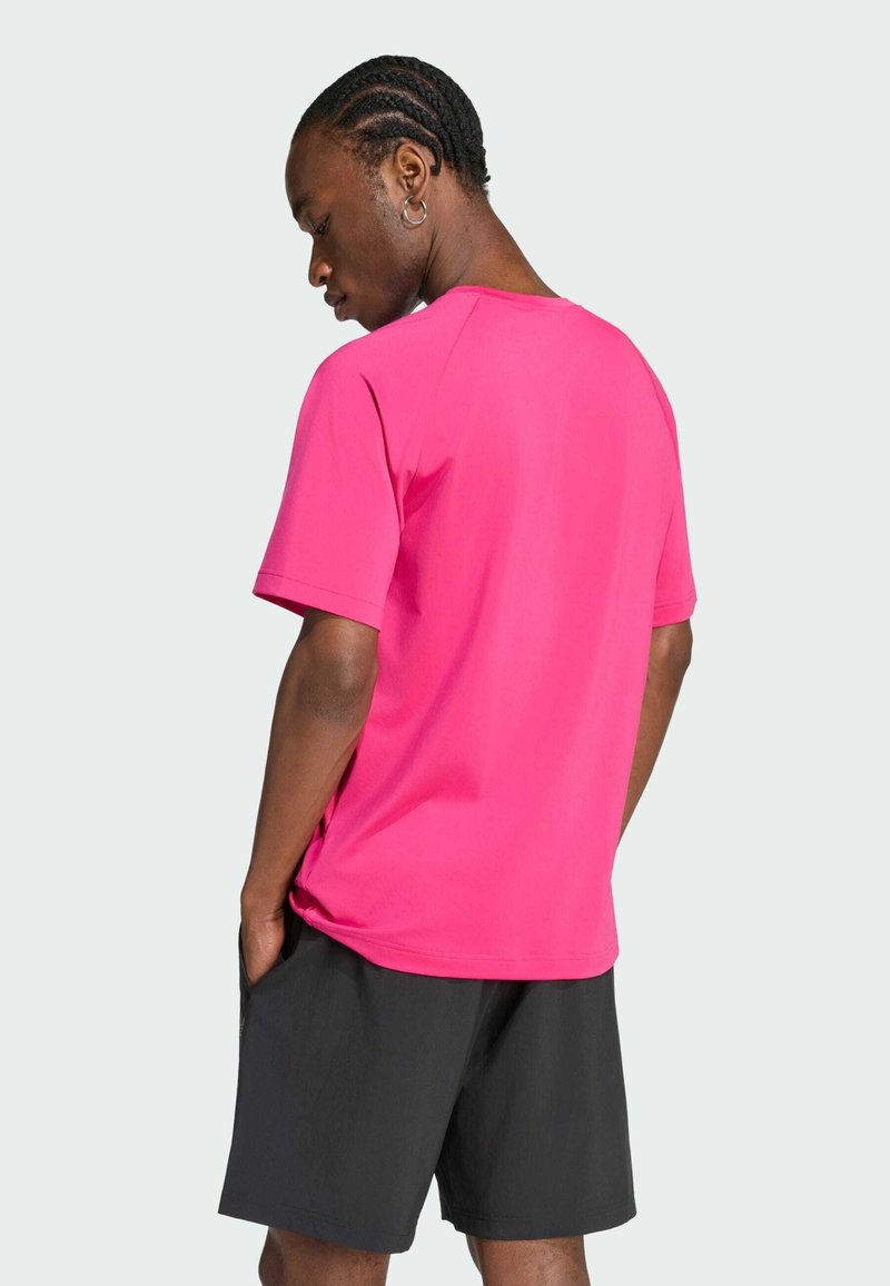 Hombre con el pelo trenzado, vistiendo una camiseta de manga corta rosa brillante y pantalones cortos negros, de pie con las manos en los bolsillos, mirando ligeramente hacia otro lado.