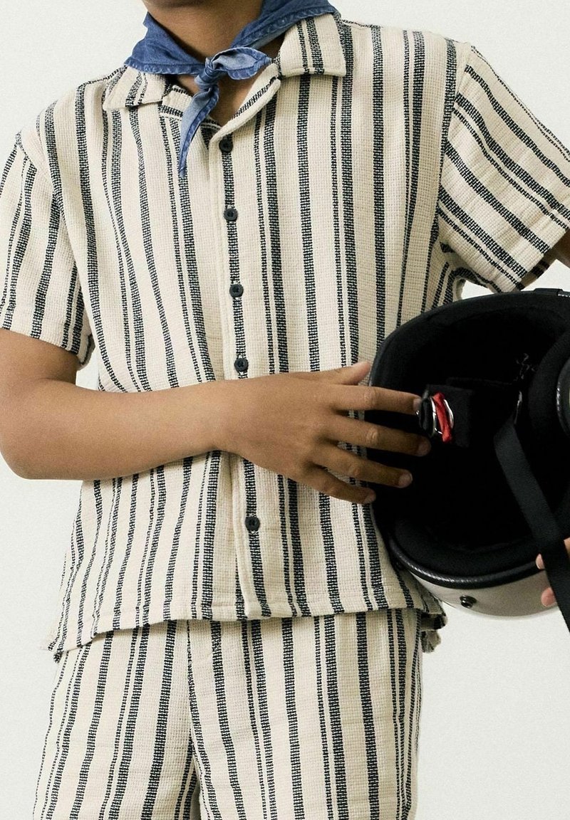 Personne portant une chemise à manches courtes beige et noire à rayures verticales et un short assorti, tenant un casque noir avec un détail de boucle rouge.