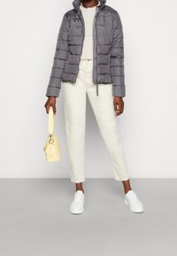 Grå dunjakke med quiltet design, lynlåslukning og høj krave. Kombineret med lyse bukser, hvide sneakers og en gul håndtaske.