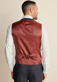 Chaleco de satén burdeos con una textura suave, que cuenta con un cierre trasero de hebilla y una correa ajustable, llevado sobre una camisa de rayas clara.