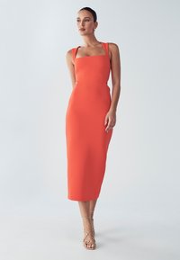 Tæt siddende midikjole i stærk orange stof med firkantet halsudskæring, brede stropper og en glat tekstur. Kombineret med guldfarvede hæle med stropper.