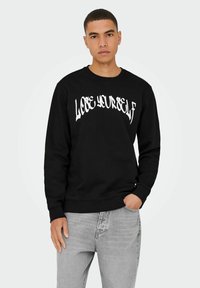 Felpa nera con testo bianco "LOOSE YOURSELF". Collo giro, maniche lunghe, polsini e orlo a coste. Indossata con jeans grigio chiaro.
