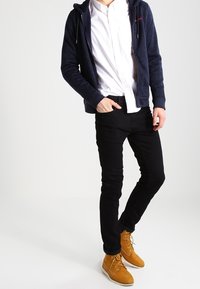 Marineblauwe zip-up hoodie over een wit overhemd met knopen, zwarte slim-fit broek en mosterdgele veterlaarzen met een gestructureerde afwerking.