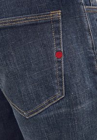 Donkerblauwe denim jeans achterzak met oranje stiksels en een klein rood rond klinknagel detail aan de zijnaad.