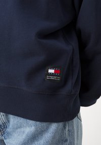 Sudadera azul marino en suave mezcla de algodón, con puños y dobladillo acanalados. Parche del logo de Tommy Jeans prominente en la parte inferior izquierda.