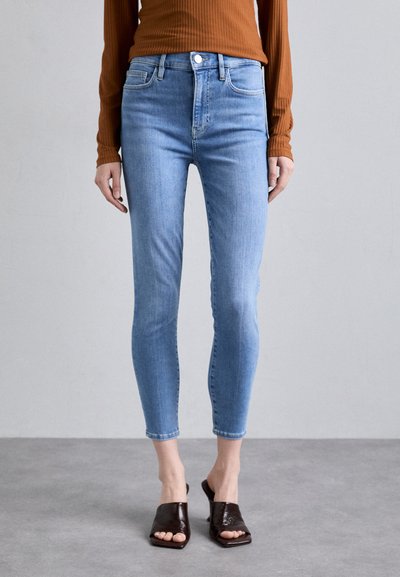Jean skinny bleu clair avec taille haute, présentant une fermeture à bouton et un léger délavage. Associé à des mules ouvertes marron.