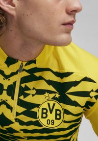 Maglia sportiva gialla e nera con un motivo geometrico, colletto a mezza zip e logo circolare con "BVB 09". Texture liscia e sintetica.