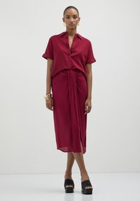 Robe chemise bordeaux à manches courtes, avec un devant à boutons et un détail de taille nouée, associée à des sandales à plateforme noires. Texture de tissu lisse.