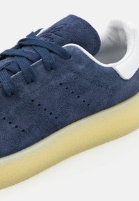 Mörkblå mockaskor med perforerad detalj, platta mörkblå snören och en ljus creamfärgad texturerad sula. Vita detaljer nära hälen.